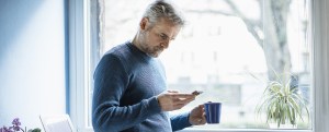Mature man standing with coffee mug standing in living room looking at smartphone
