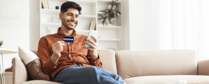 Fast Online Shopping. Smiling young Arab guy holding debit credit card in hand and using cell phone, making financial transaction sitting on the couch at home in living room, free copy space