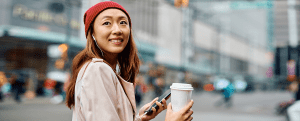 Happy Asian woman using smart phone while carrying takeaway coffee and walking on the street