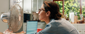 woman using a laptop, leaning towards a fan to cool down.